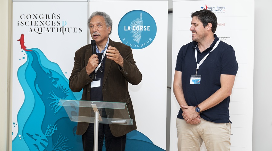 Daniel Pauly and Fabrice Teletchea, co-organizers of the International Congress on Aquatic Sciences in Saint Pierre and Miquelon. talking in front of the blue and white congress banner